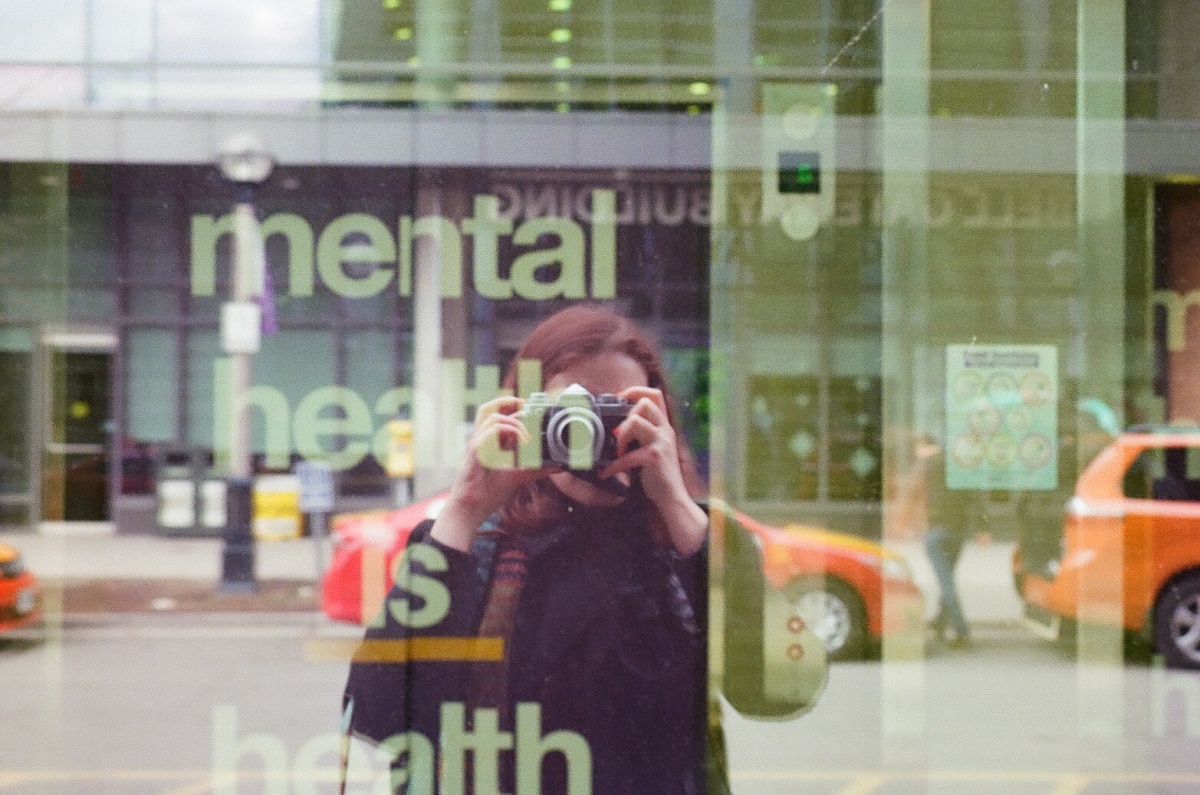 Person looking through a camera looking at her reflection in the street in a window that reads Mental health is health.