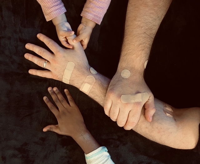 On a black background we see a pair of male adult arms, the right hand holding onto the left arm, the man is wearing a wedding ring on the left hand, and several plasters covering the arms. A pair of toddler's hands hold on to the left thumb, and another child's hand rests next to the left hand.